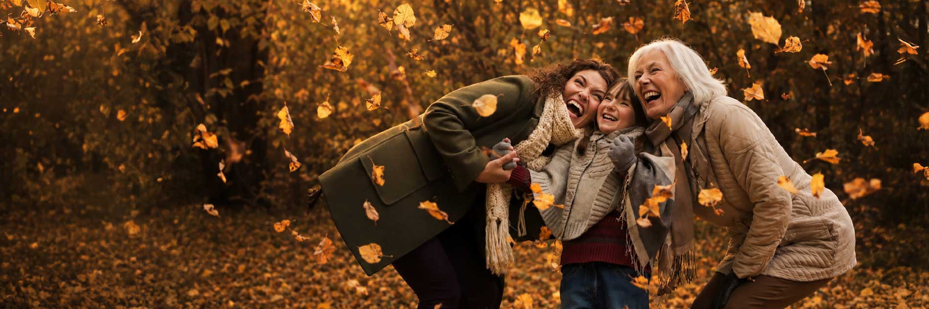 Three generations of women laughing together outdoors as autumn leaves fall around them